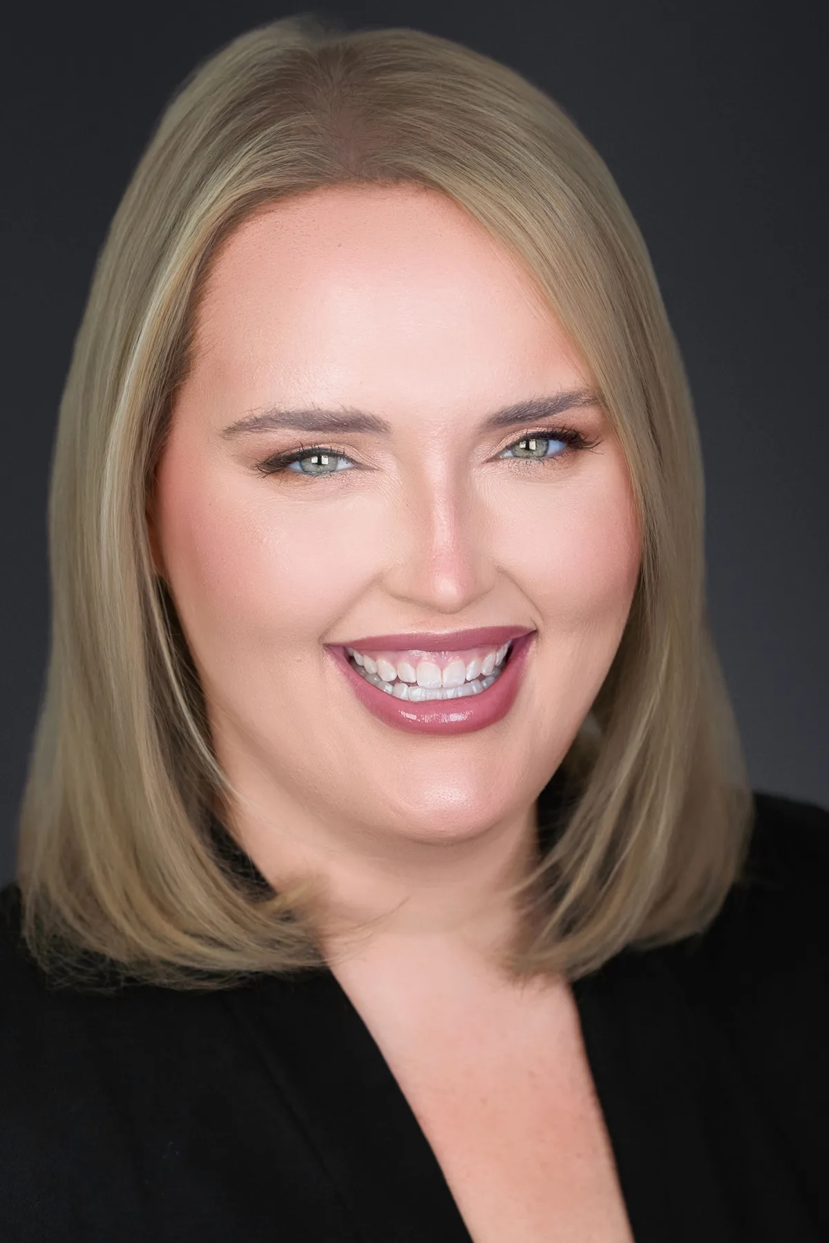 A woman with a blonde bob in a black top, smiling, on a charcoal backdrop.