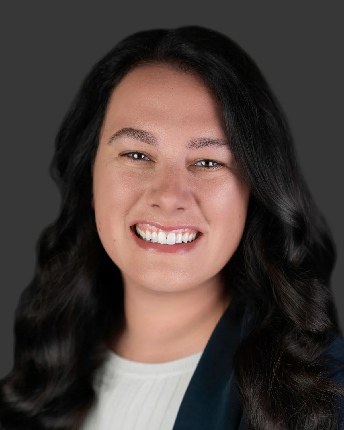 A woman with shoulder-length brown hair in a camel blazer, smiling, on a slate backdrop.