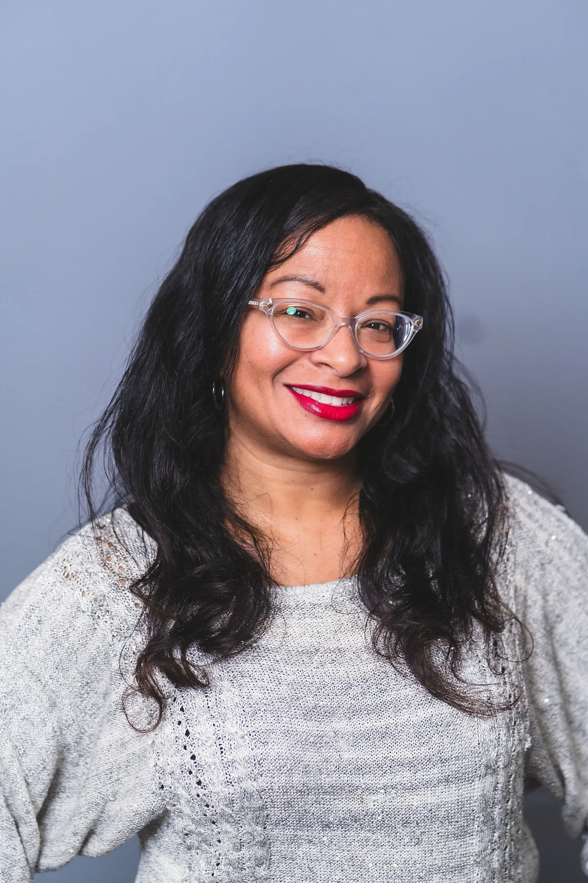 A woman with long dark hair and clear-framed glasses in a gray sweater, smiling, on a pale blue-gray backdrop.