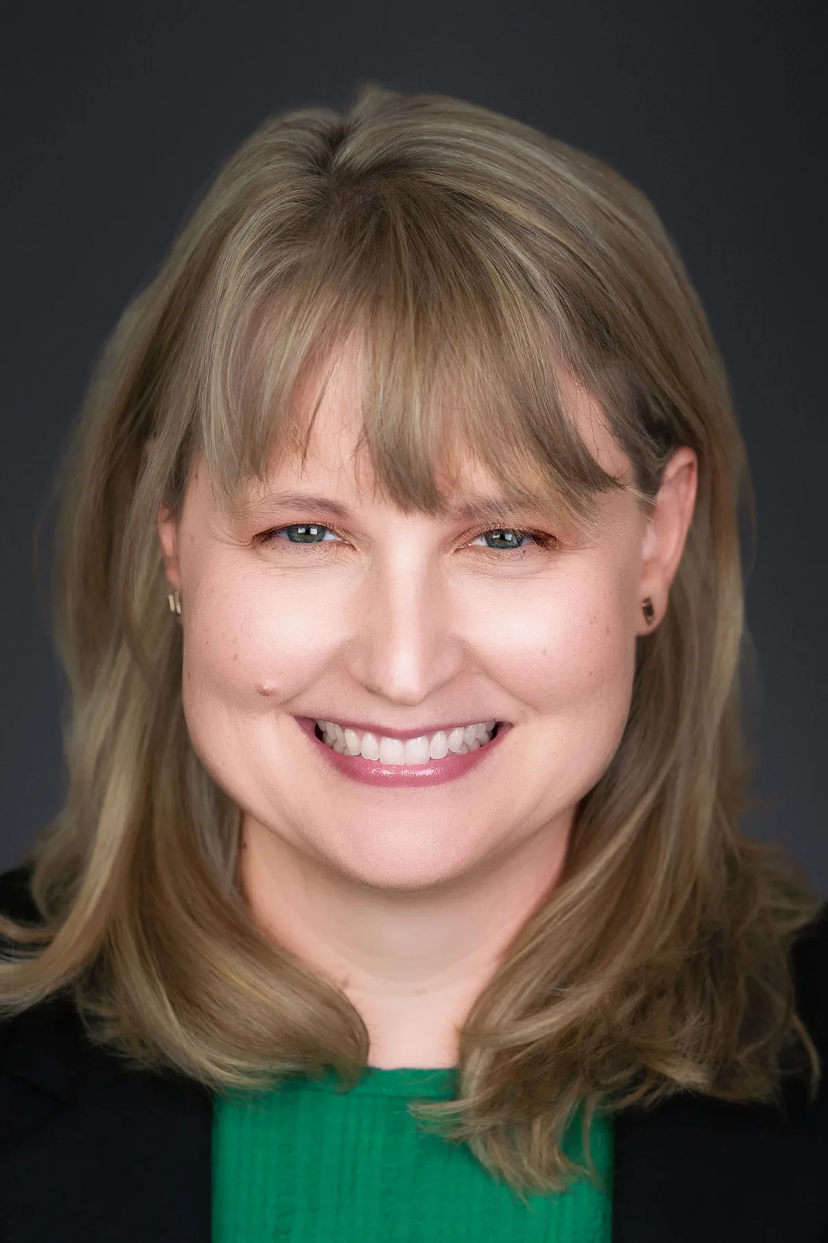A woman with shoulder-length hair and bangs in a black blazer and green top, smiling, on a charcoal backdrop.