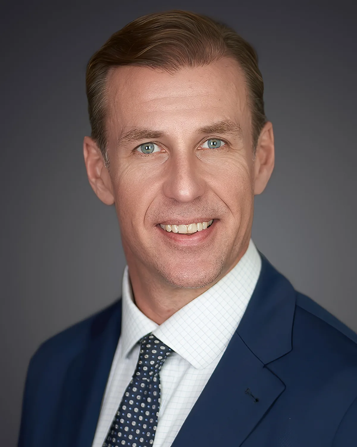 A man in a navy suit, checked shirt and dotted tie, smiling, on a gray backdrop.
