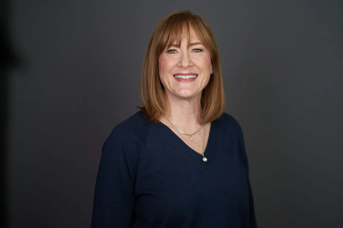 A woman with an auburn bob in a navy sweater and a pendant necklace, smiling, on a charcoal backdrop.