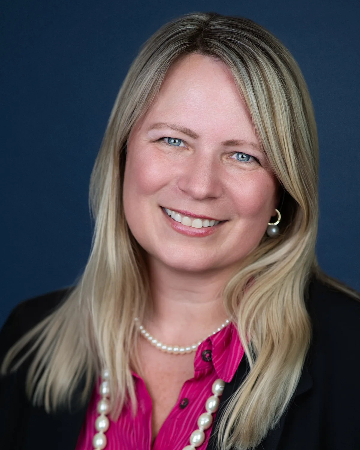 A woman with long blonde hair, pearls and a pink blouse, smiling, on a navy backdrop.