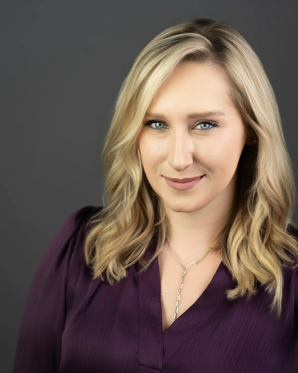 A woman with wavy blonde hair in a plum blouse, a slight smile, on a gray backdrop.