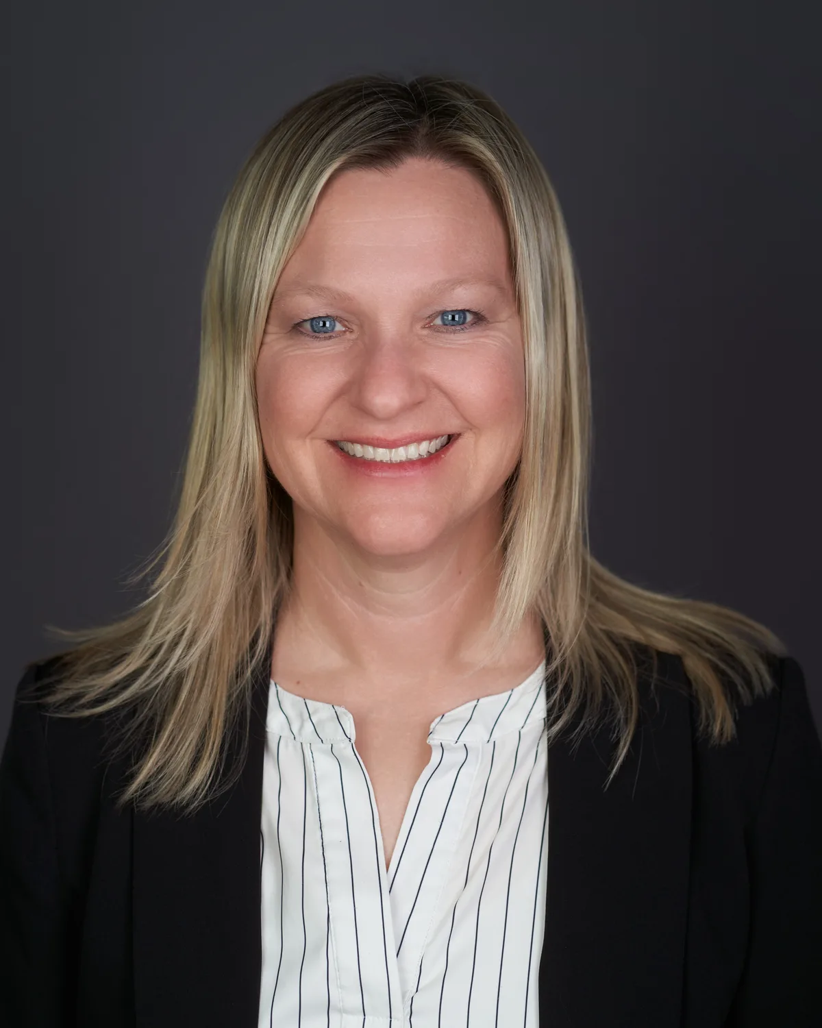 A woman with straight blonde hair in a black blazer and striped blouse, smiling, on a charcoal backdrop.