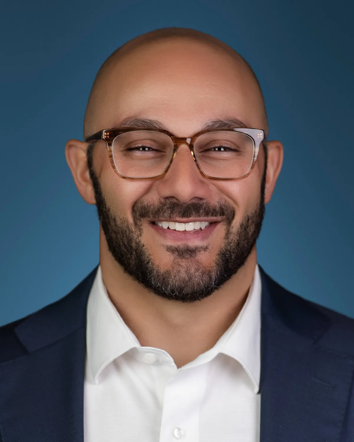 A bearded man in glasses, navy suit and white shirt, grinning, on a deep blue backdrop.
