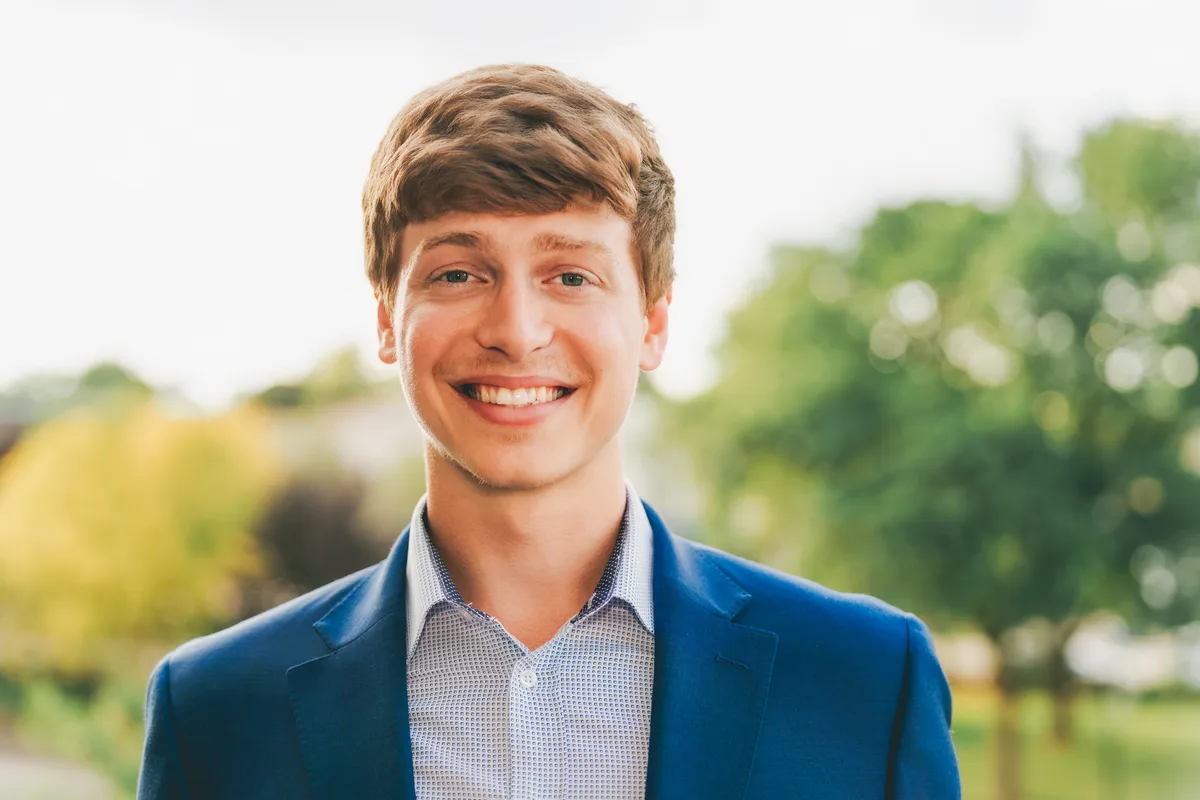 A young man in a blue blazer grinning in open shade, green trees soft behind him.