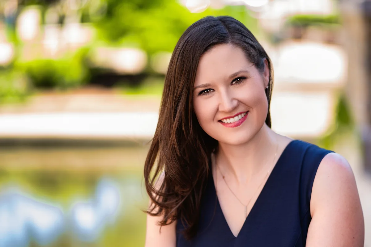 A woman with long dark hair in a navy sleeveless top, smiling, the riverwalk soft and green behind her.