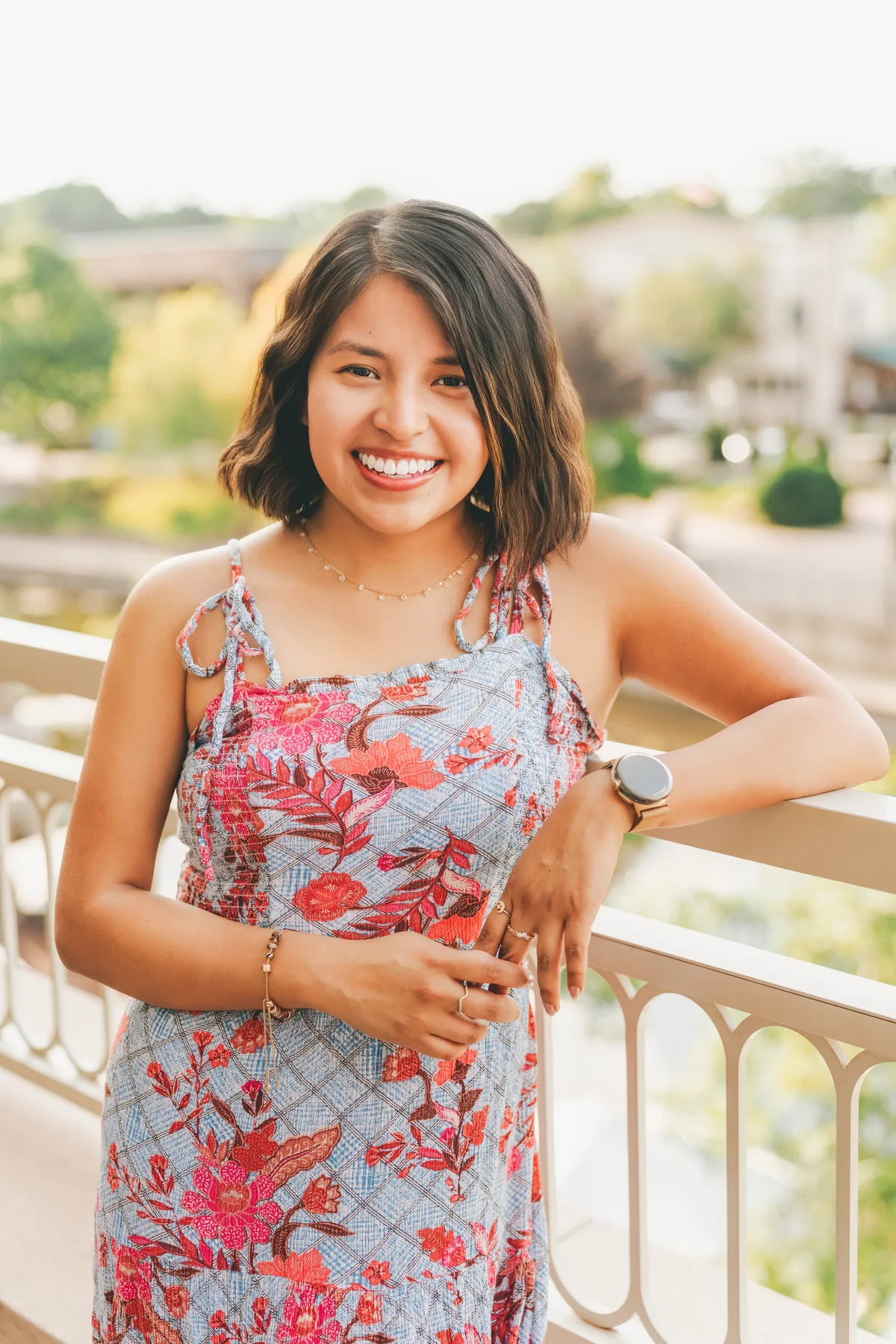 A smiling woman in a floral dress leaning on a white railing, a riverwalk soft behind her.