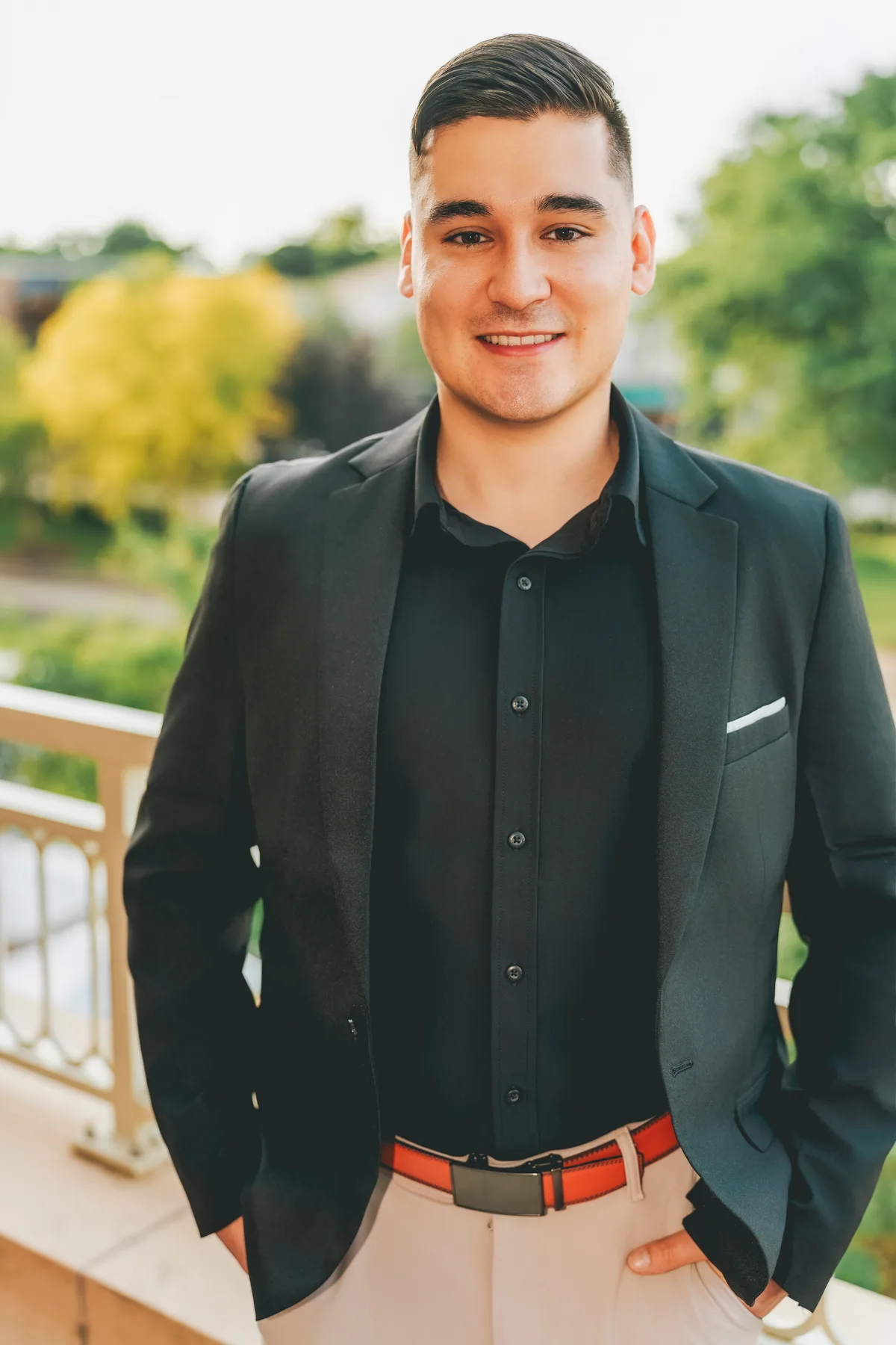 A man in a black blazer, hands in pockets, smiling at a riverwalk railing with trees behind.
