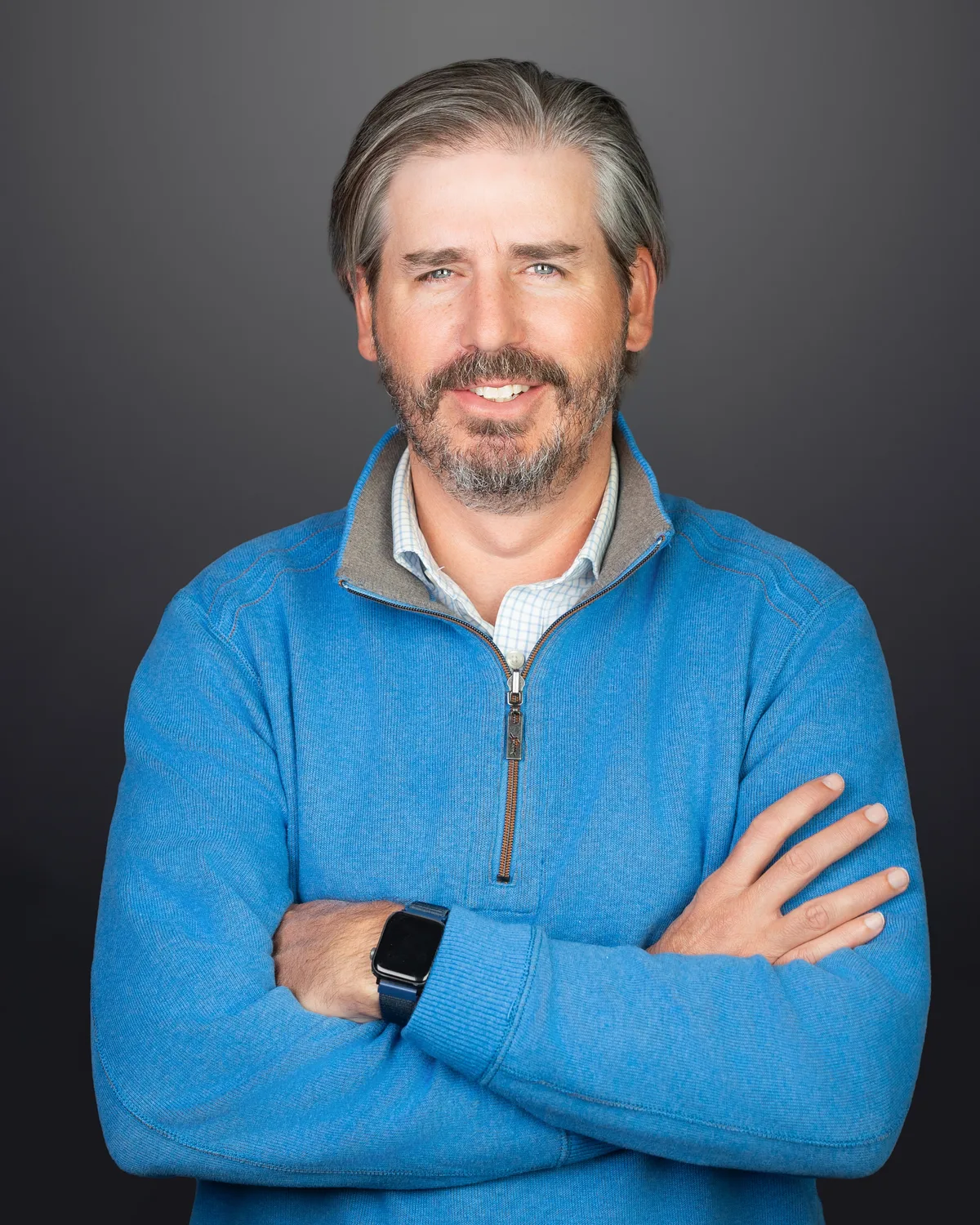 A man in a bright blue quarter-zip, arms crossed, smiling on a dark gray backdrop.