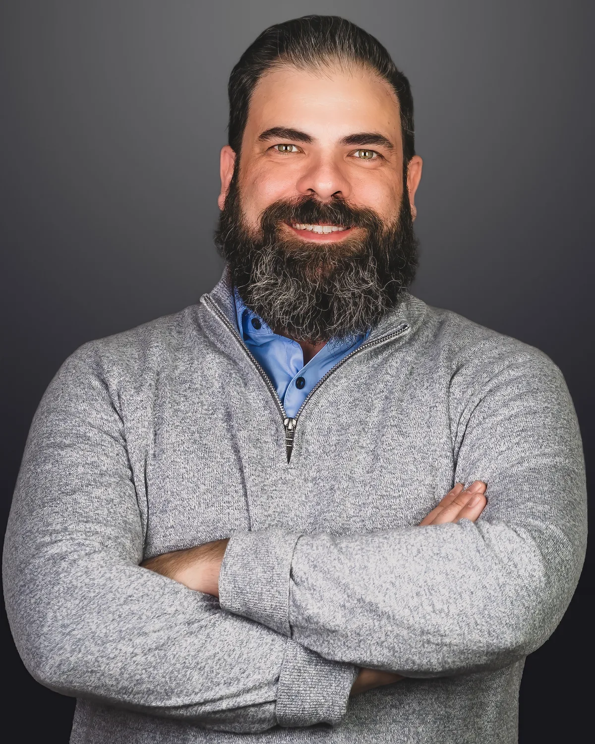 A bearded man in a gray quarter-zip, arms crossed, smiling on a dark gray backdrop.