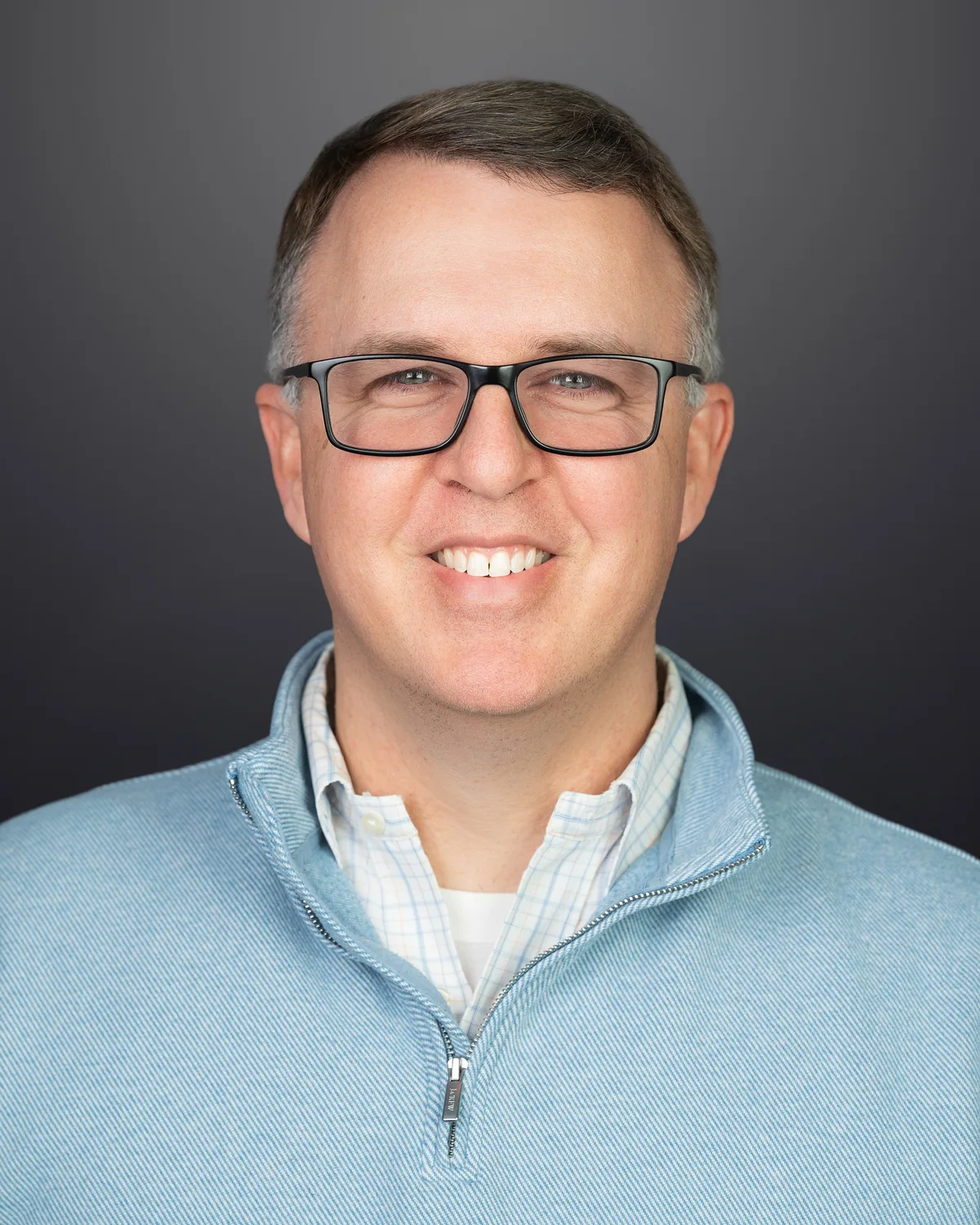 A man in glasses and a light blue quarter-zip, smiling, on a dark gray backdrop.