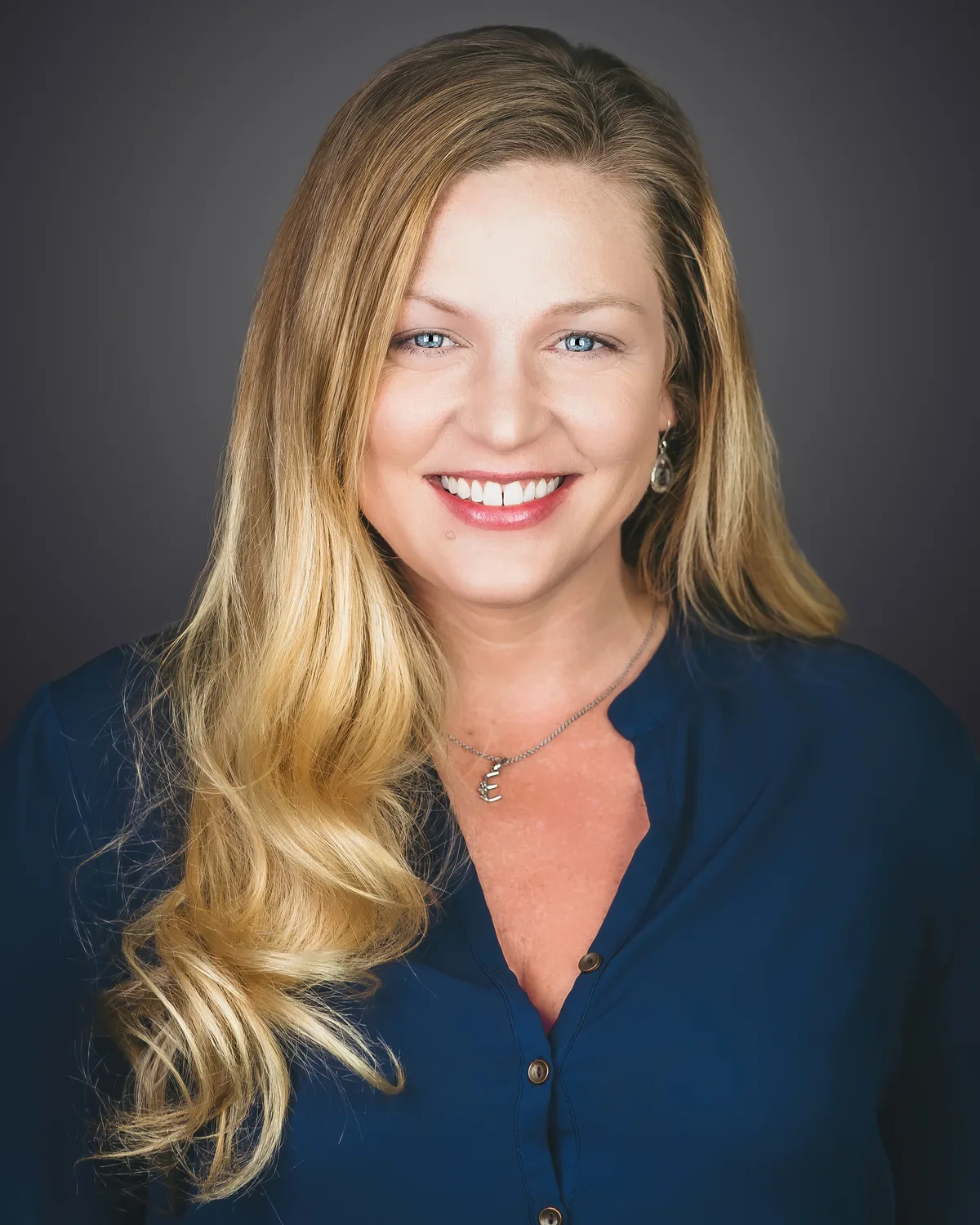 A woman with long blonde hair in a navy blouse, smiling, on a dark gray backdrop.