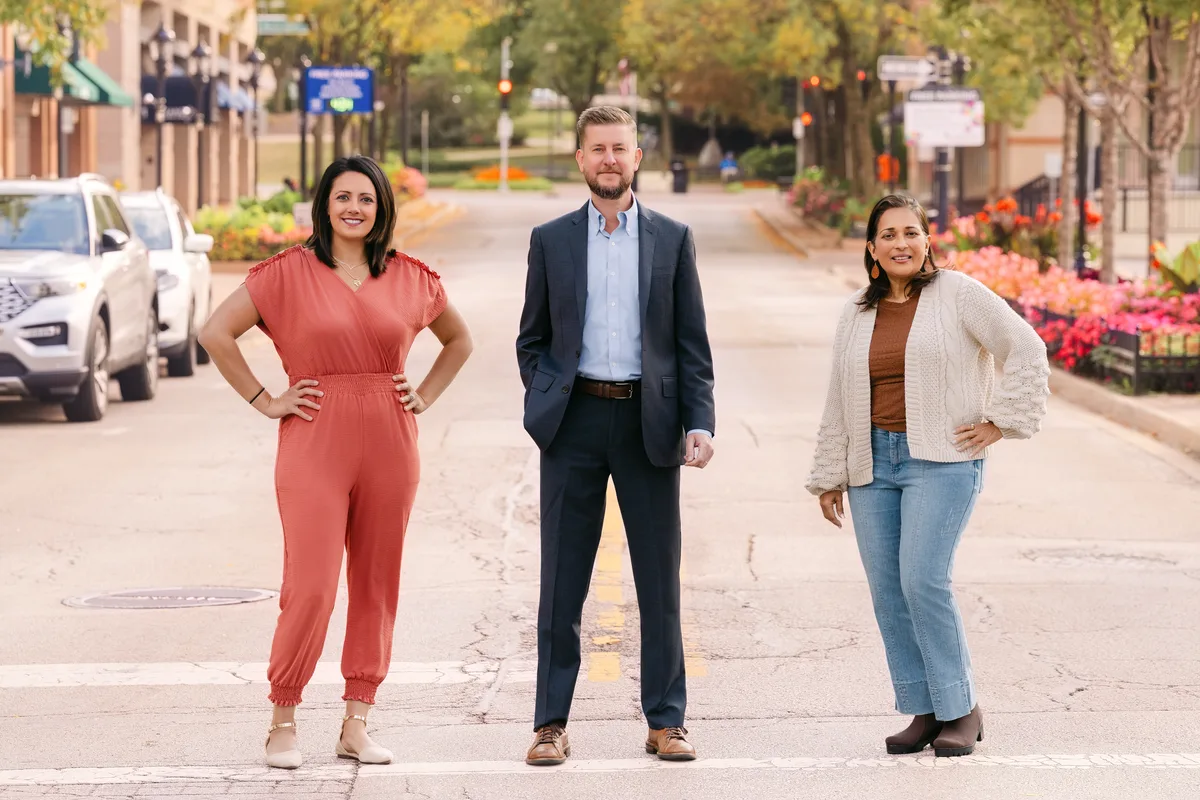 A team of three standing in a crosswalk on a tree-lined downtown street, flower beds and storefronts behind them.