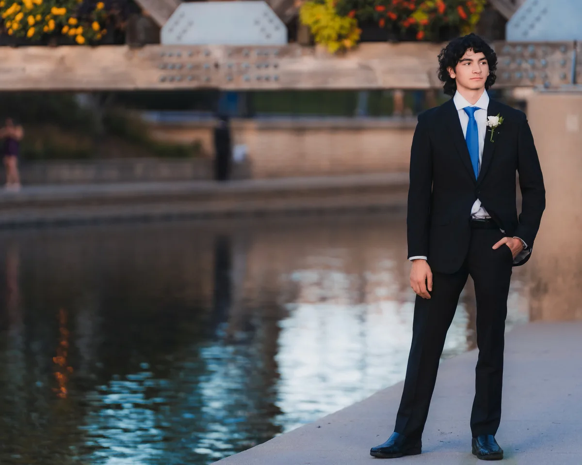 A high school senior in a black suit and blue tie stands by the water on the Naperville Riverwalk at dusk.