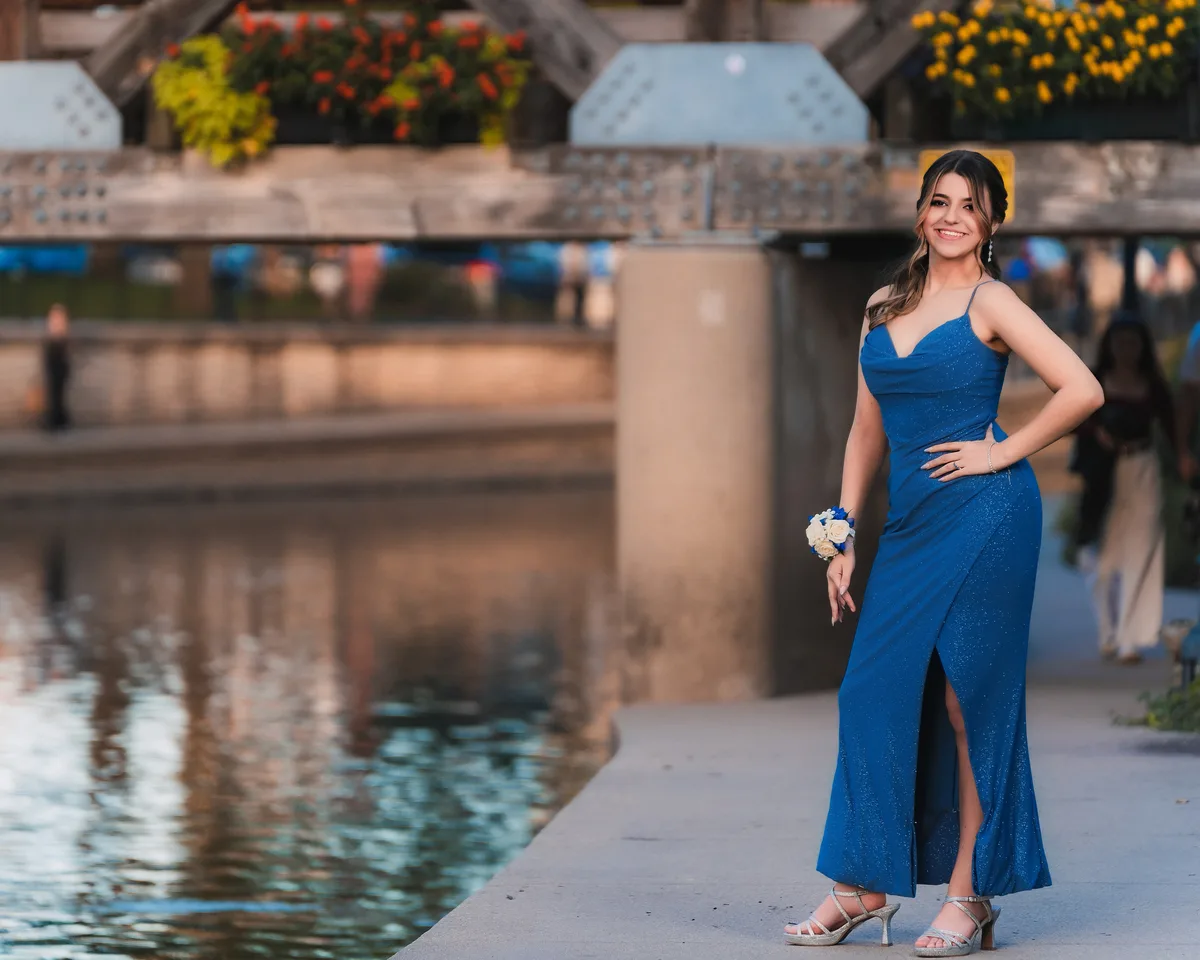 A high school senior in a long blue gown stands on the Naperville Riverwalk, the covered bridge and evening water behind.