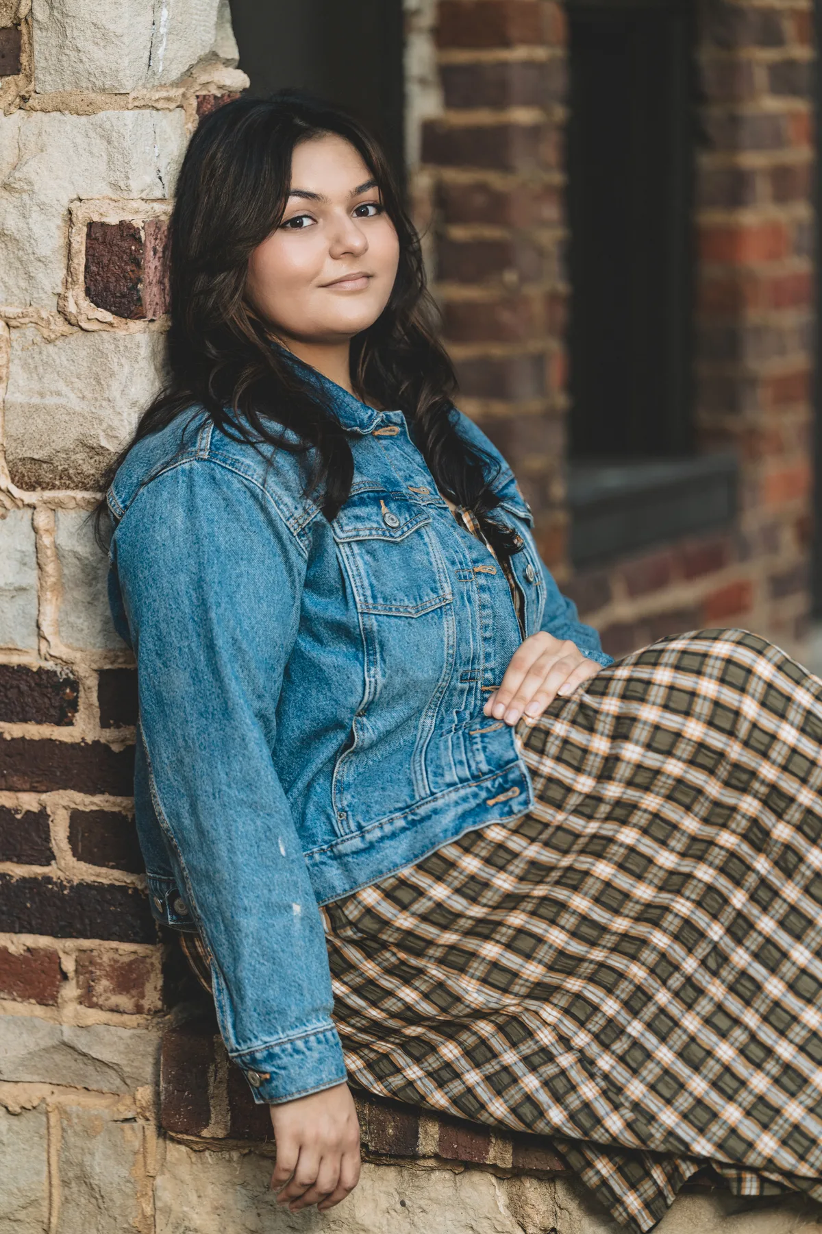 A high school senior leans back against a brick wall, seated, in a denim jacket and long plaid skirt.