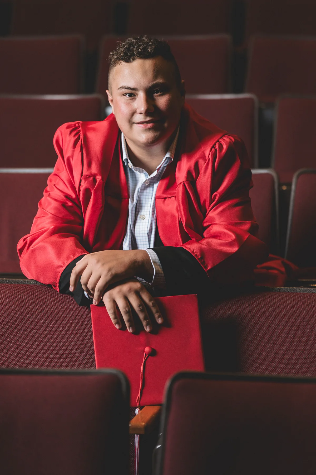 A high school senior in a red graduation gown leans on a row of auditorium seats, cap in hand.