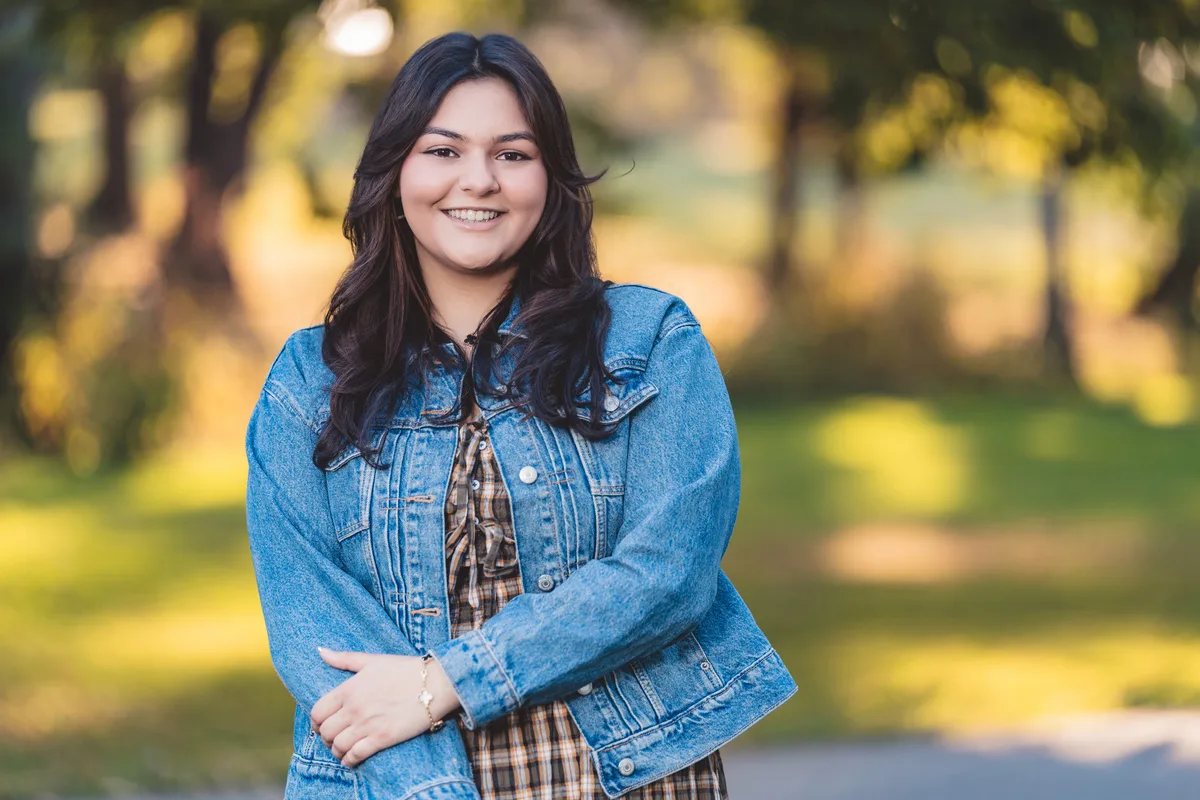 A high school senior in a denim jacket stands on a park path, arms crossed, fall color behind.