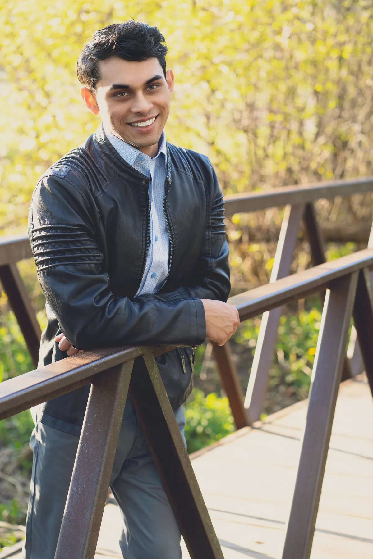 A high school senior in a leather jacket leans on the rail of a park footbridge, spring green behind.