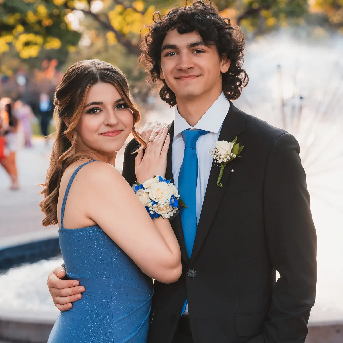A homecoming couple in a blue dress and black suit, a fountain lit by evening sun behind them.