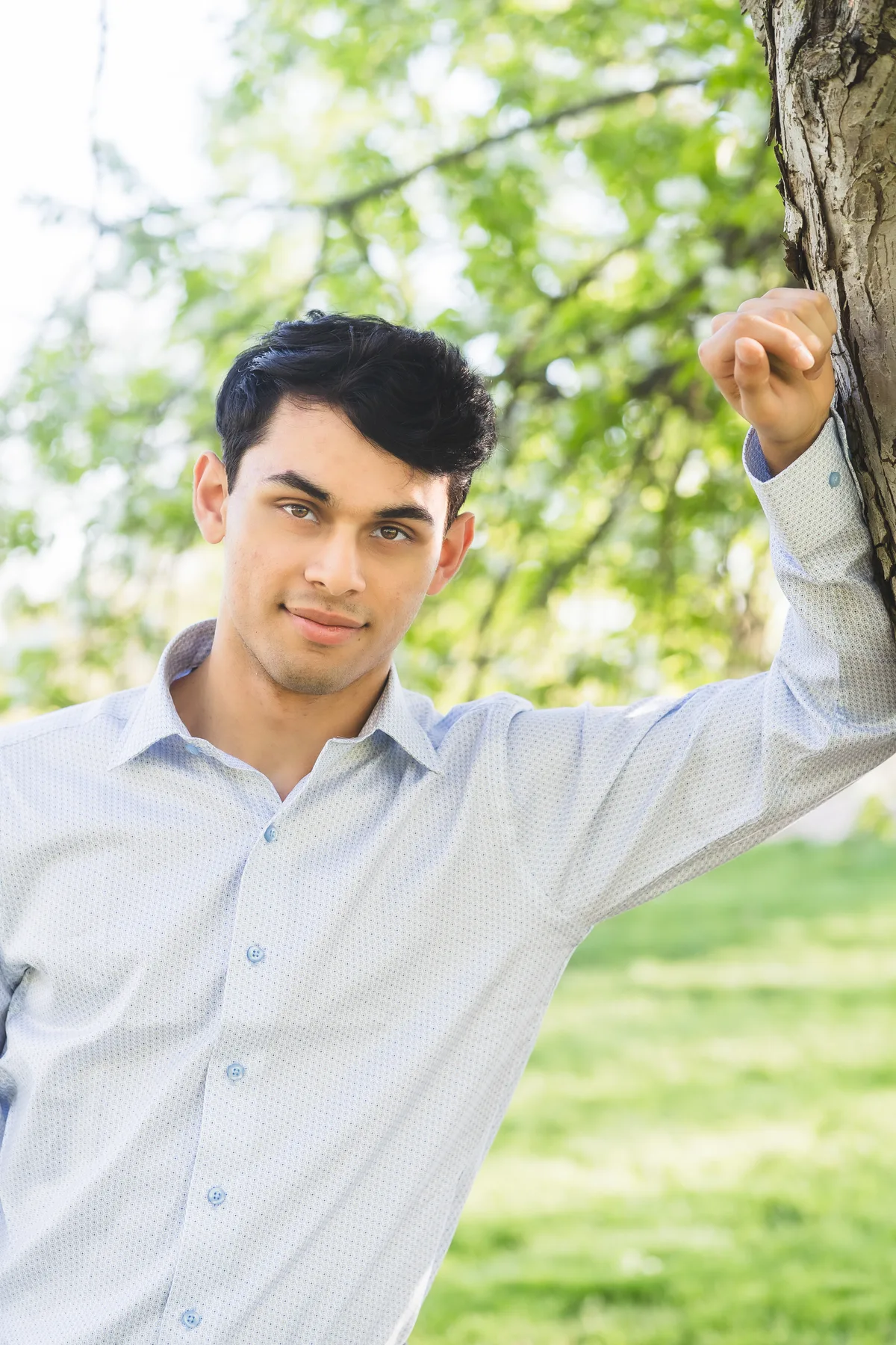 A high school senior rests a hand on a tree trunk in a park, spring leaves behind.
