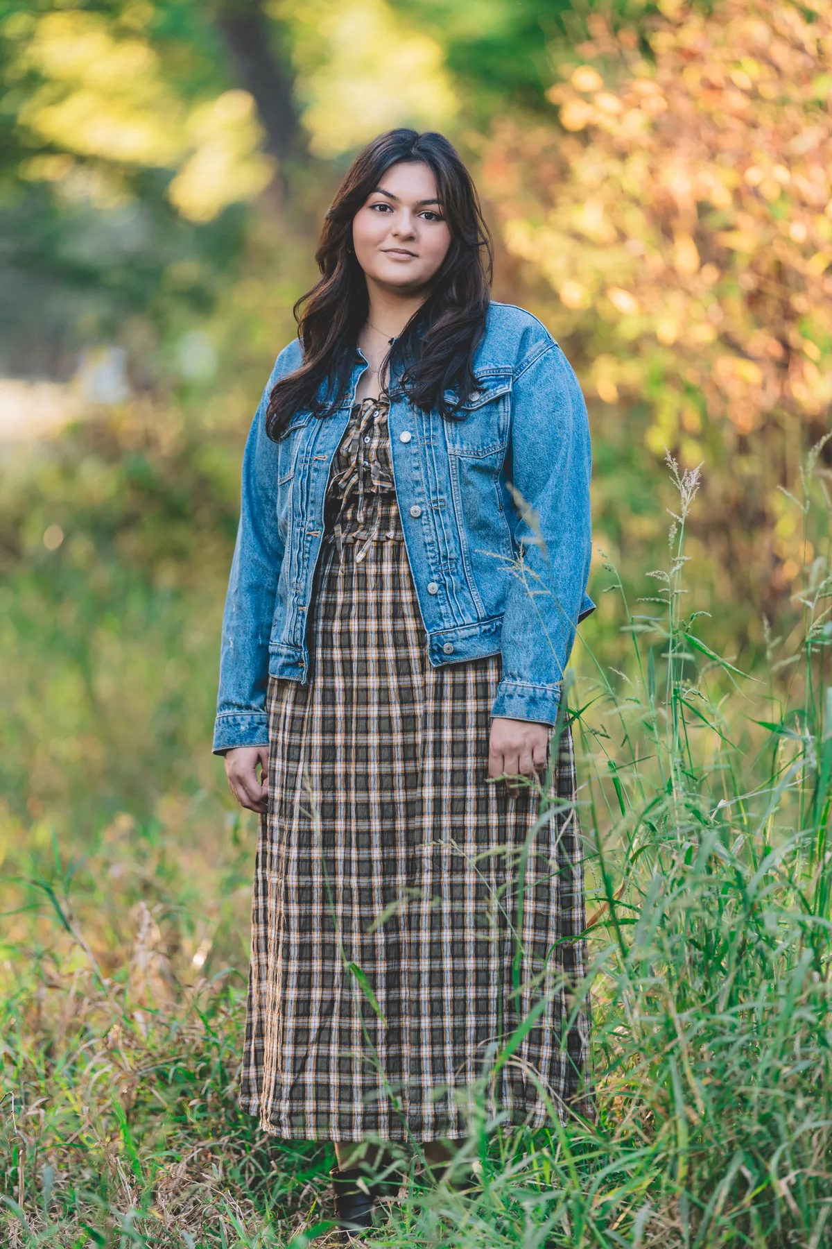 A high school senior stands in tall grass in an open field, fall color behind.