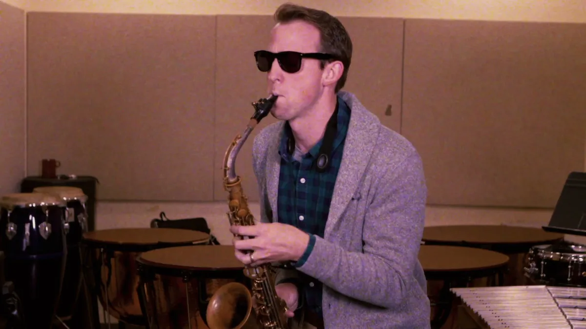 A man in sunglasses and a gray cardigan plays alto saxophone in a percussion room, timpani and congas behind him.