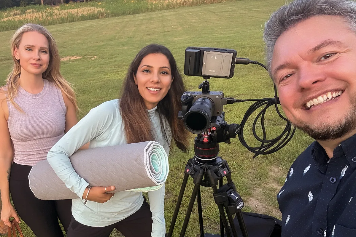 Greg Mulvey takes a selfie on set with two of the Washoga team, one holding a rolled yoga mat, beside a camera on a tripod.