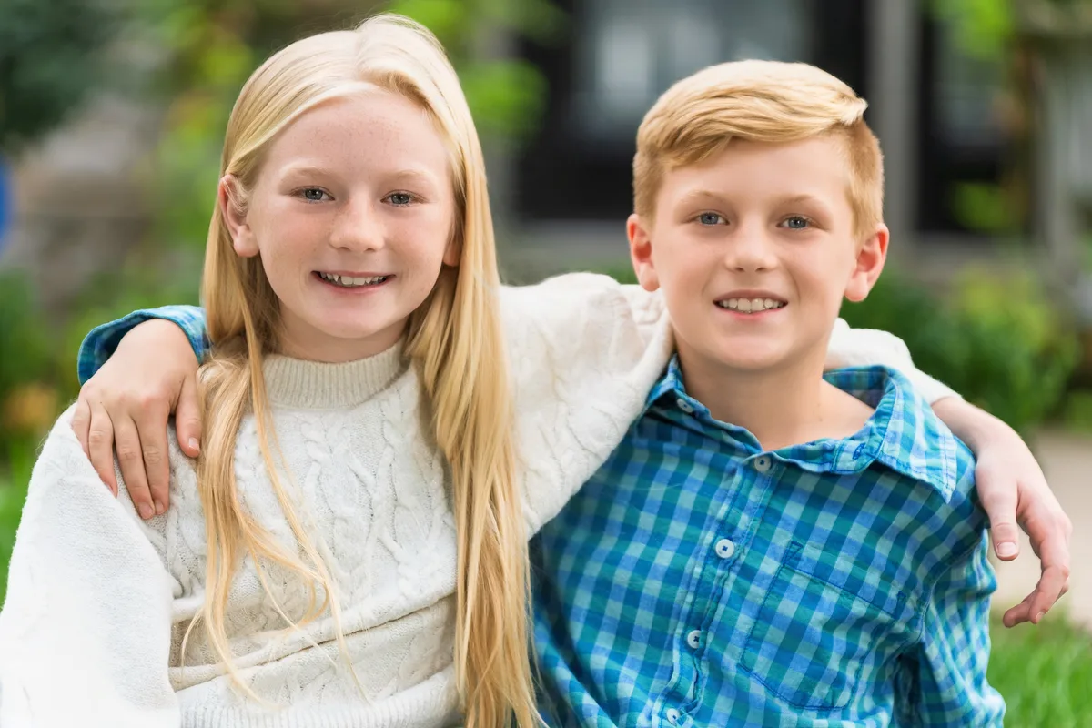 A brother and sister side by side in their backyard, her arm around his shoulder.