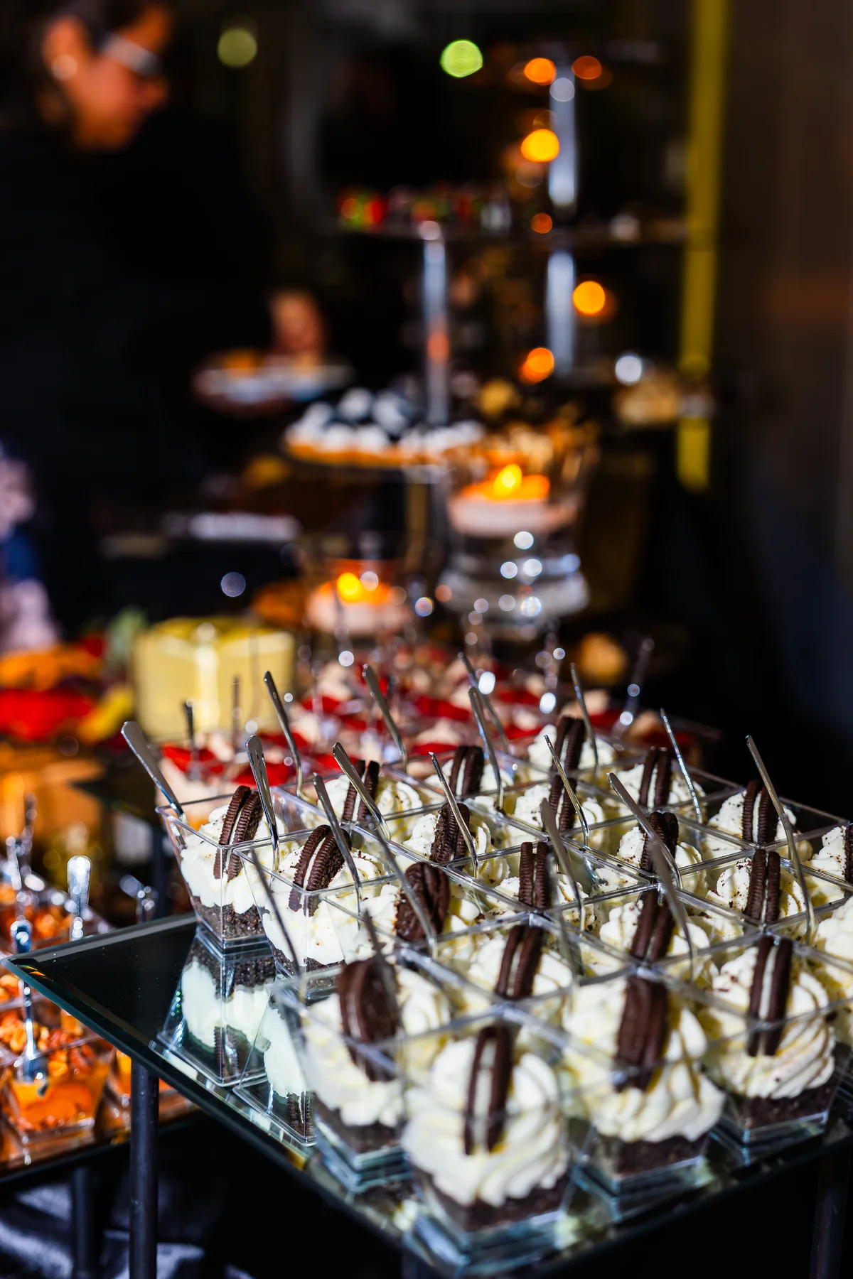 Rows of cream dessert cups topped with chocolate sandwich cookies on a mirrored tray, a candlelit table blurred behind.