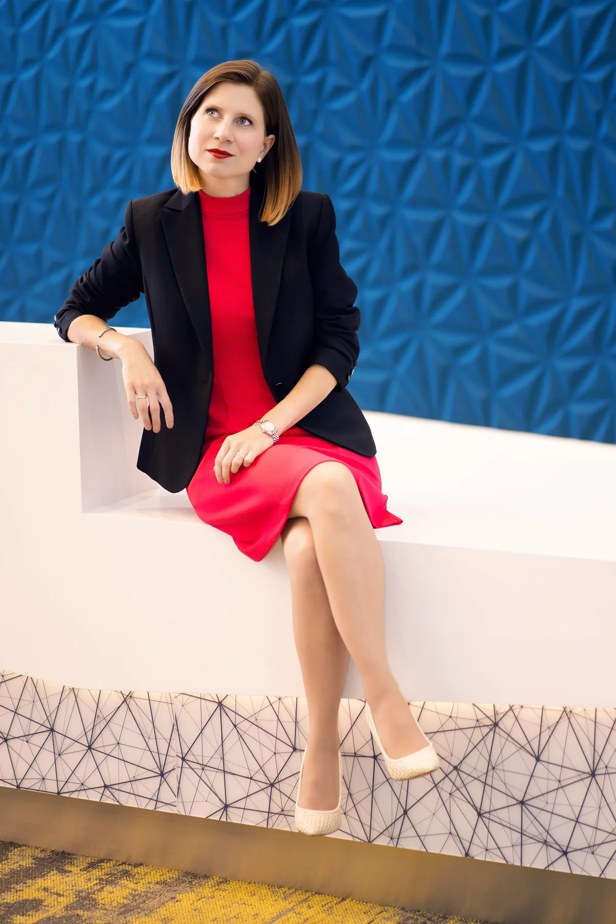 A headshot made on location, seated on a low white ledge against a sculpted blue wall.