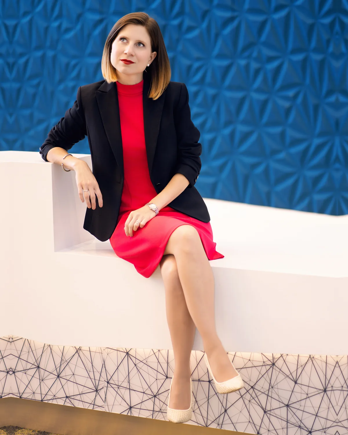 A headshot made on location, seated on a low white ledge against a sculpted blue wall.