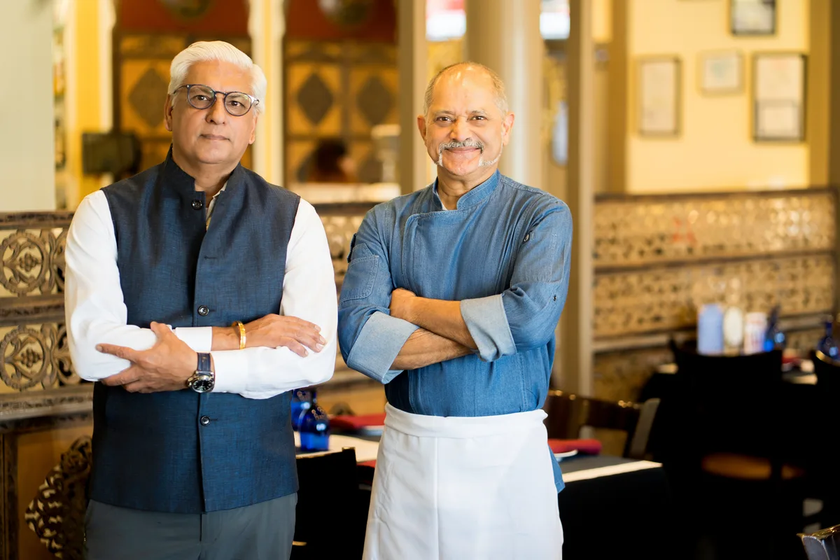 Two restaurant owners stand together in their dining room, tables set behind them.