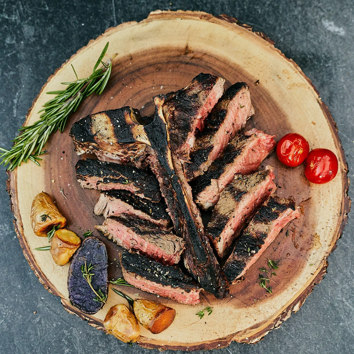 Sliced steak fanned on a wooden board with roast vegetables and herbs.