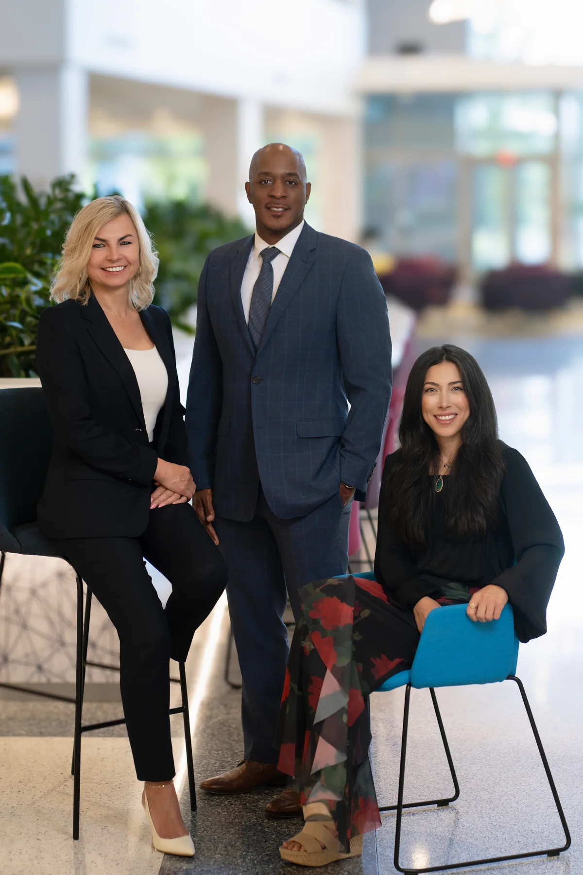 Three colleagues in their office lobby, two seated on stools and one standing between them.