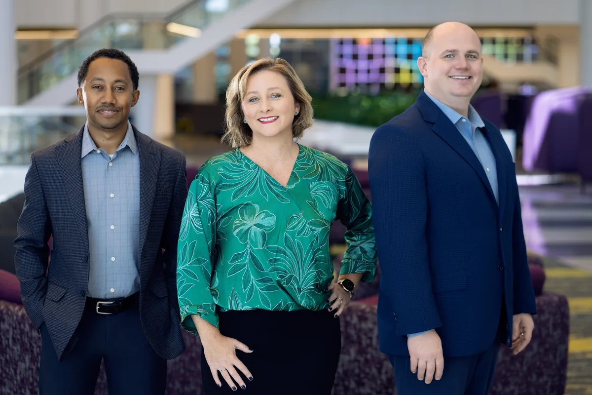 Three colleagues standing together in a corporate lobby, a staircase and glass behind them.