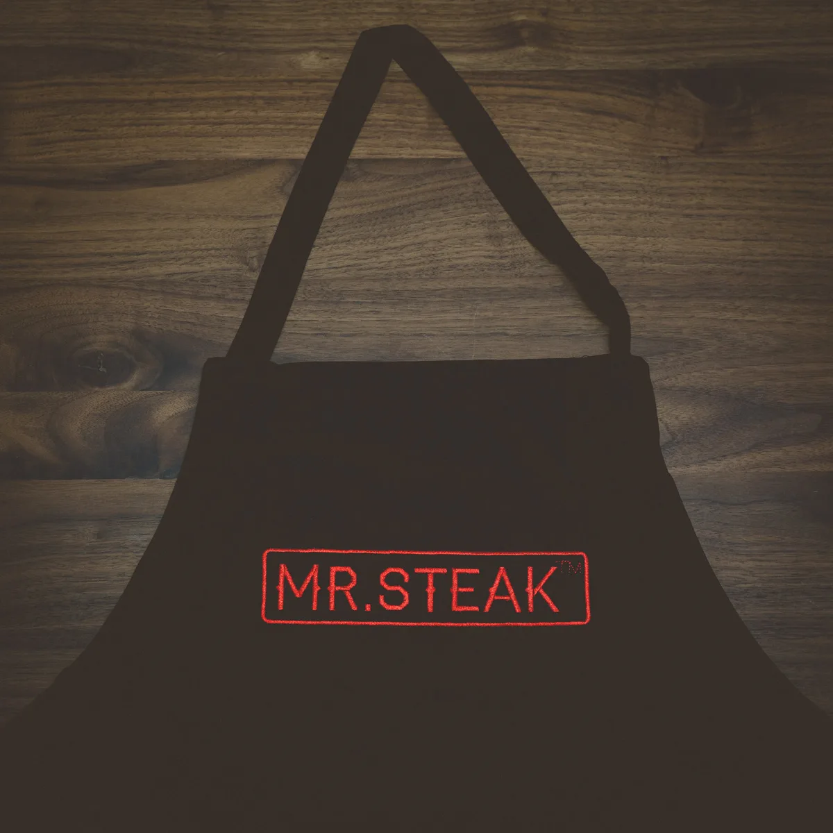 A black Mr. Steak apron with the red logo, against dark wood.