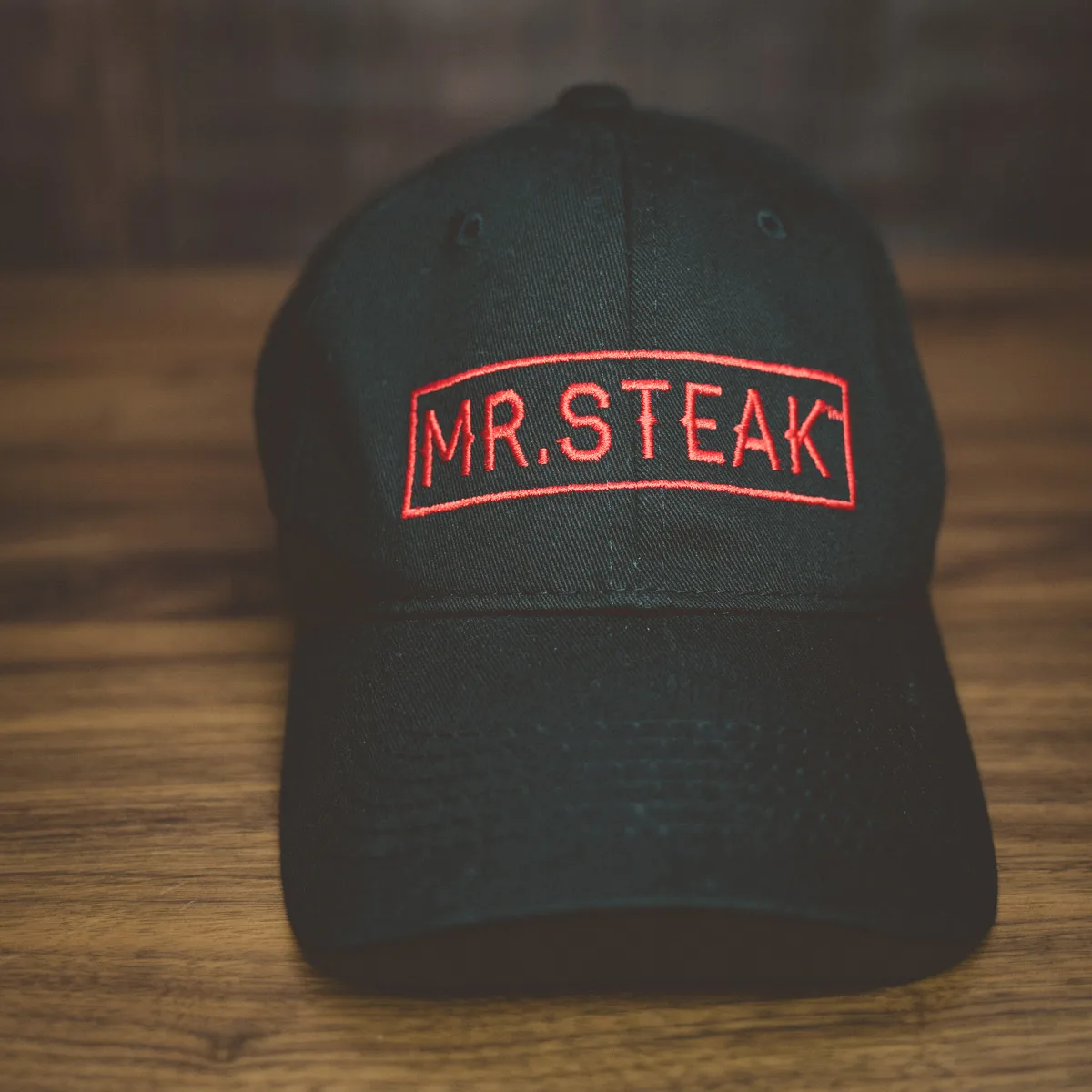 A black Mr. Steak cap with the red logo on a wooden surface.