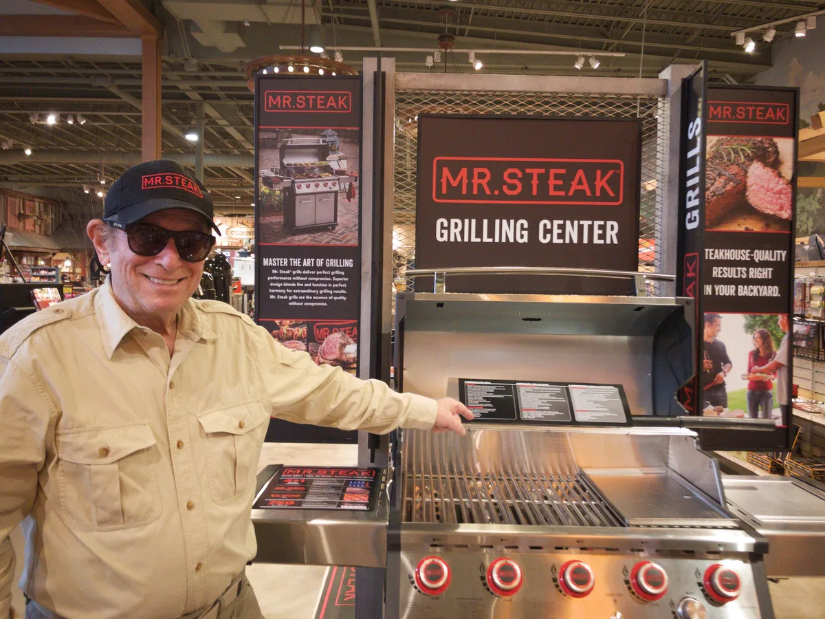 Mr. Steak's founder beside a stainless five-burner grill under a Mr. Steak Grilling Center display at Bass Pro Shops.