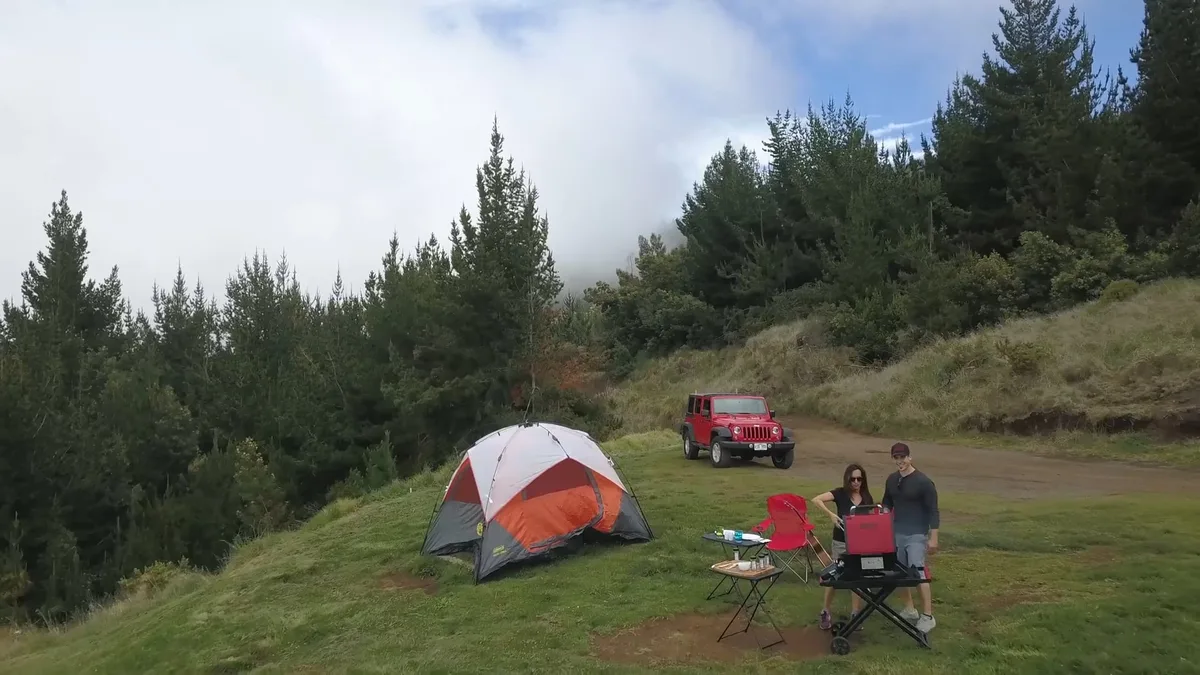 Opening frame of the portable grills spot: a tent, a red Jeep and two campers at a grill on a forested hillside.