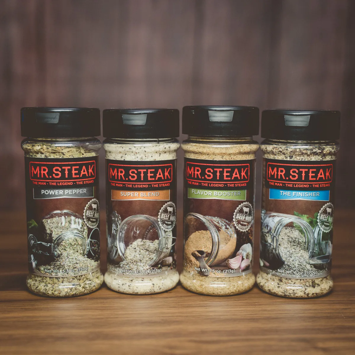 Four Mr. Steak seasoning shakers lined up on a wooden surface.